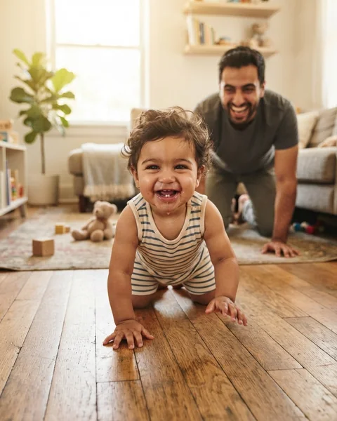 An eight-month-old baby crawling straight at the camera with a huge grin while his father laughs behind him